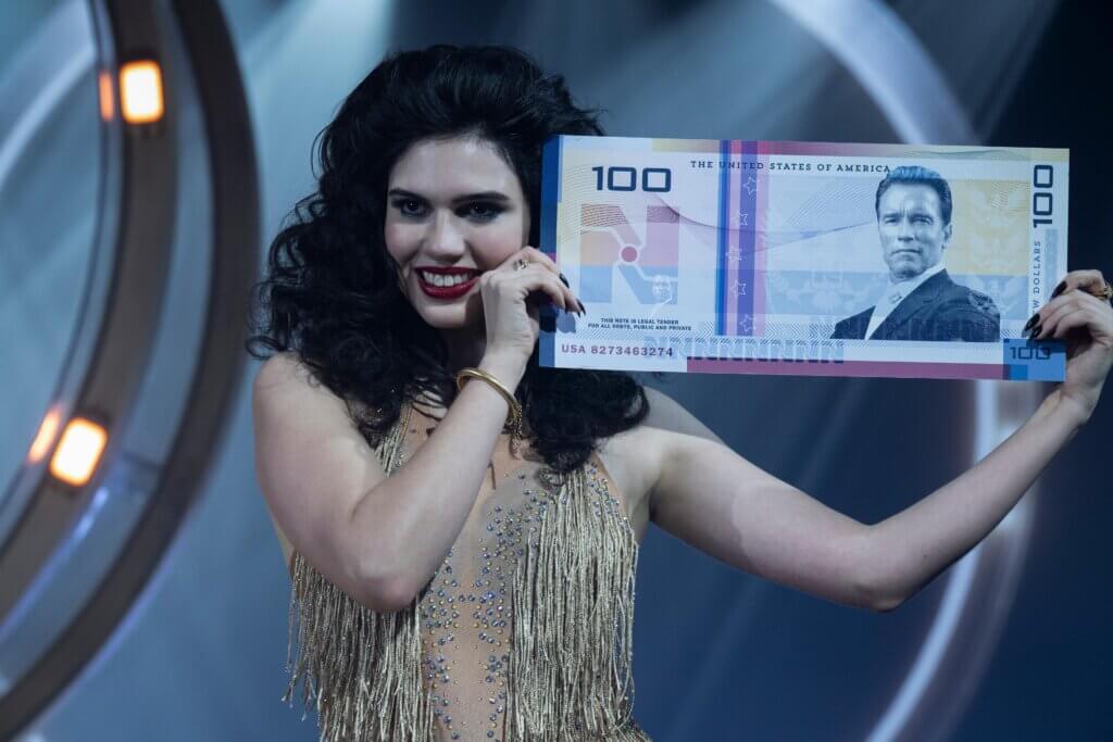 una azafata del concurso sosteniendo un gran billete de Nuevos Dólares con la cara de Arnold Schwarzenegger como referencia a la anterior adaptación de 1987.
